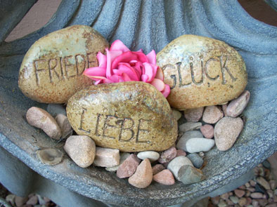 Drei Steine mit den Wörtern Frieden, Glück und Liebe, umgeben von kleinen Steinen und Blüten.