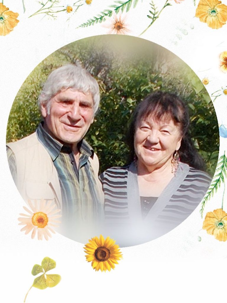 Ein älteres Paar steht lächelnd vor einer grünen Kulisse, umrahmt von Blumen.