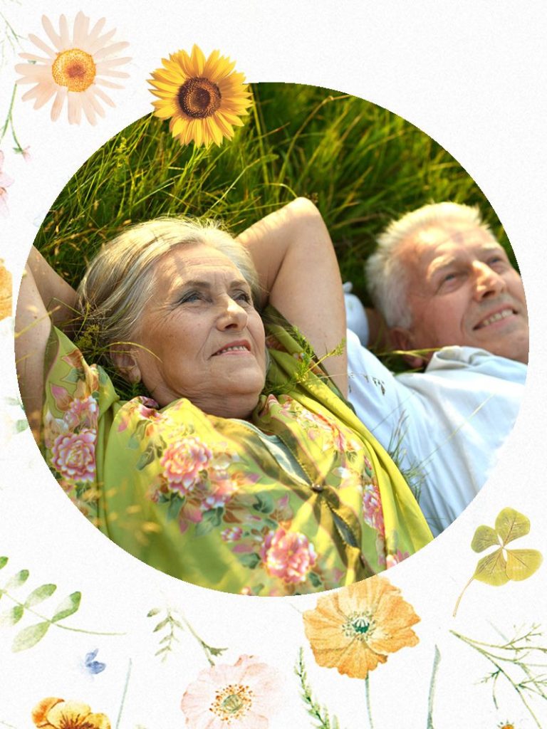 Zwei ältere Personen liegen in der Wiese und genießen die Sonne, umgeben von Blumen.